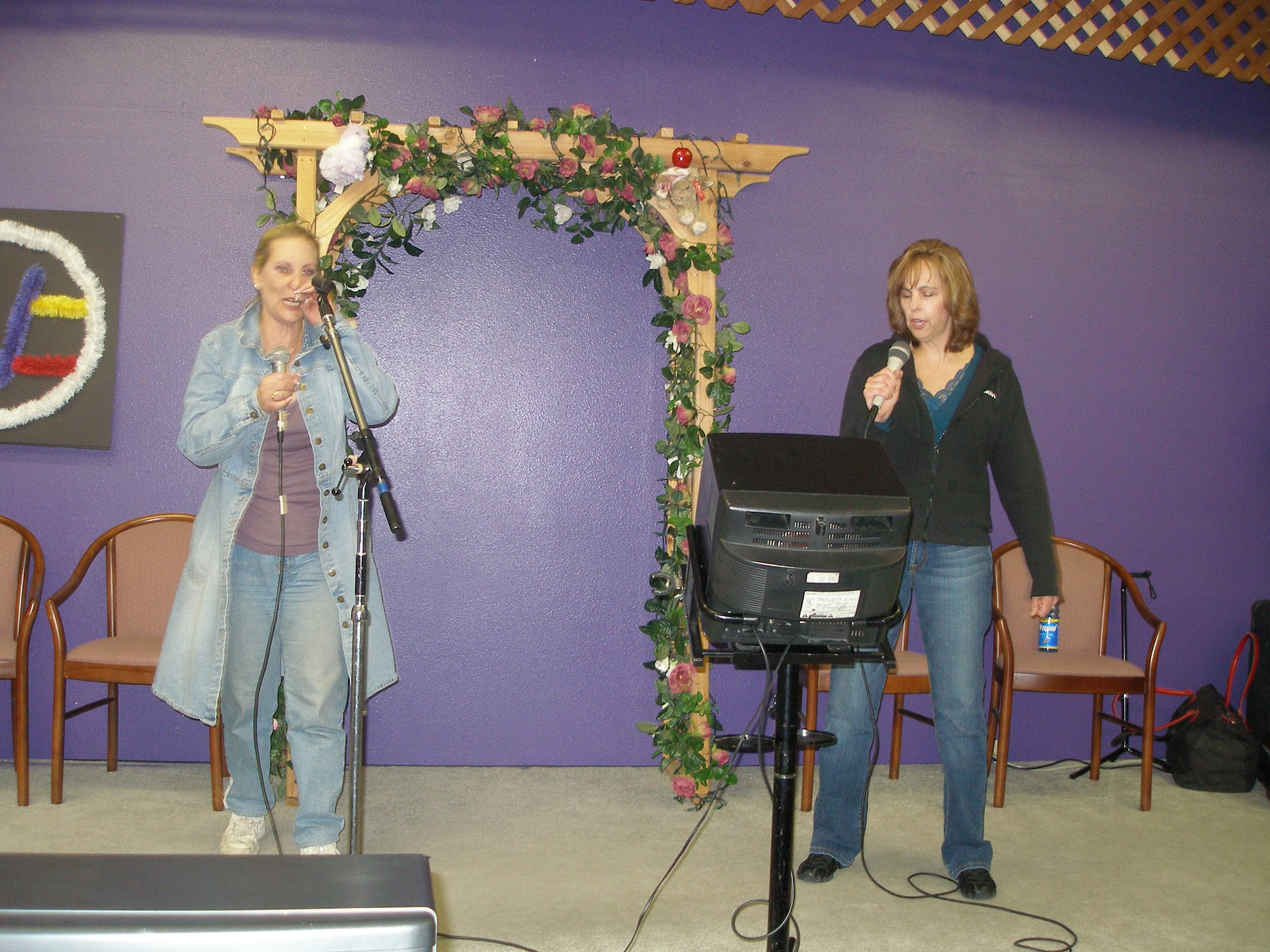 Two women singing together on the Oasis CSL stage with floral arch backdrop