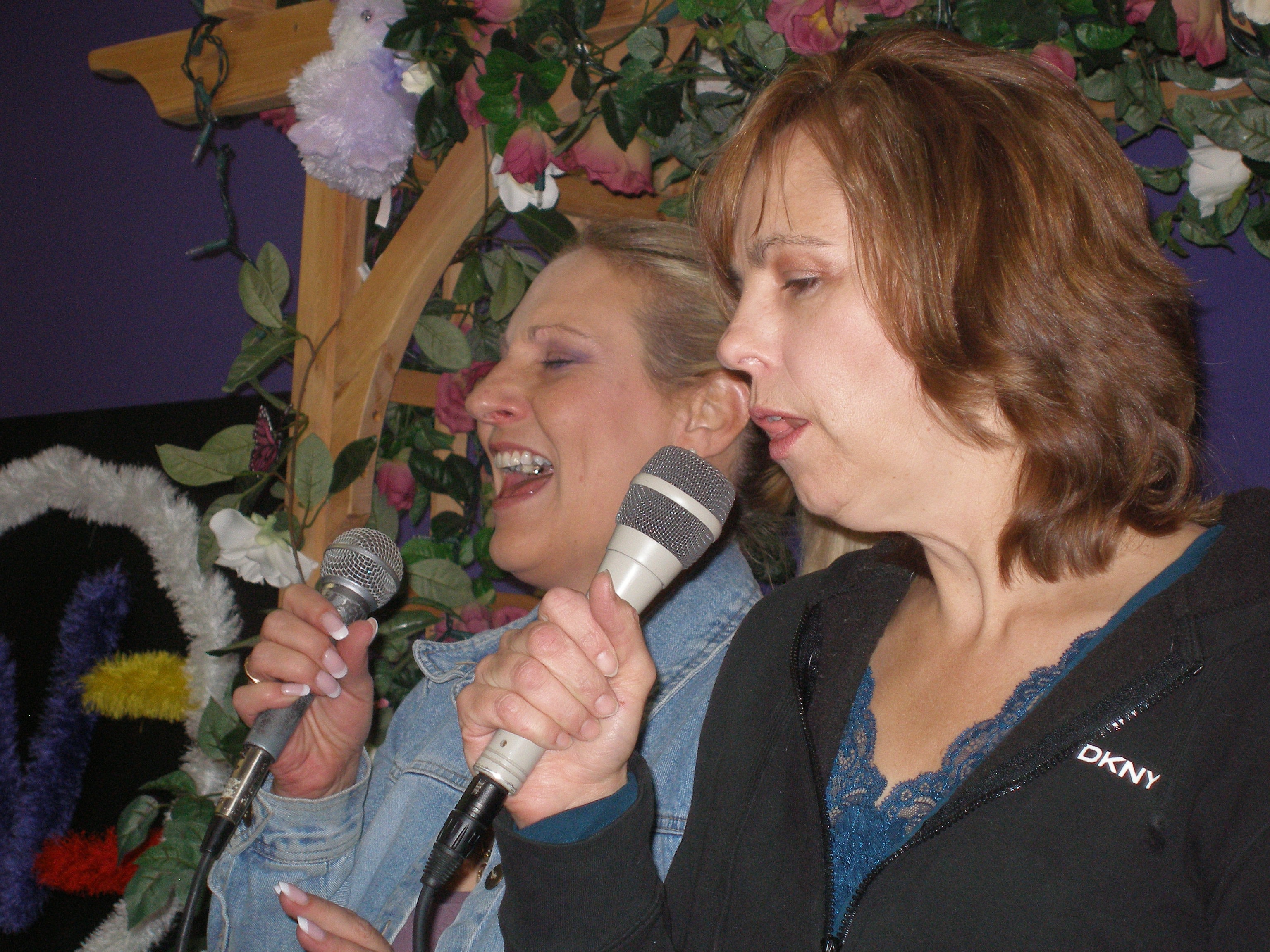 Two women singing joyfully side by side with microphones at Oasis CSL
